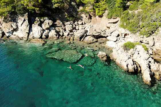 Klart turkisblåt vand ved en klippefuld kyst, hvor personer snorkler nær overfladen. (AI-genereret tekst)