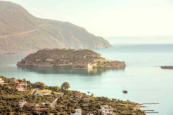 En udsigt over en historisk ø med ruiner og omkringliggende hav, og i forgrunden ses en kystby med bygninger. (AI-genereret tekst)