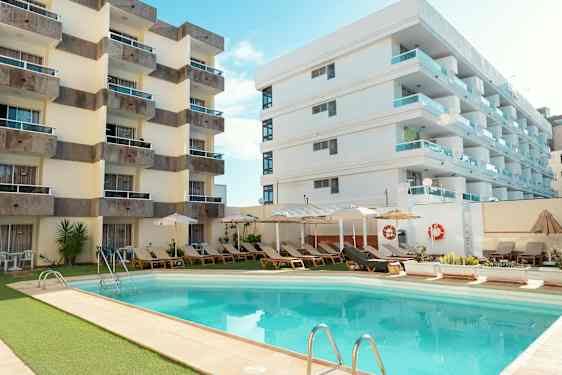 Hotelbygninger ved en udendørspool omgivet af liggestole og parasoller. (AI-genereret tekst)