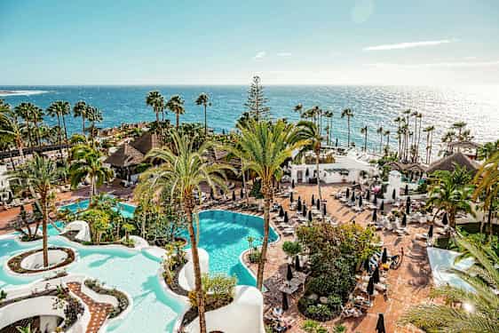 Udsigt over et hotelområde med to pooler, palmer og solsenge ved havet. (AI-genereret tekst)