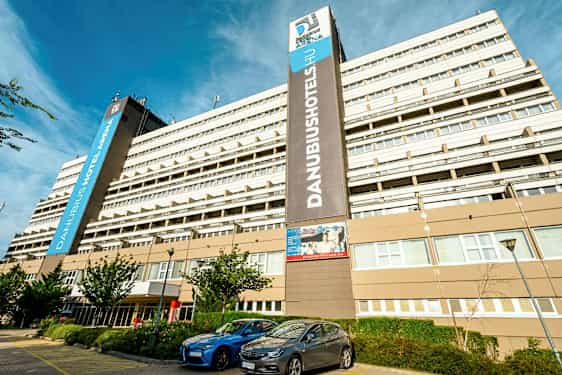 En stor hotelbygning med flere etager med skiltet ”Danubius Hotel Arena” synligt. Foran bygningen er parkerede biler og nogle små træer. (AI-genereret tekst)