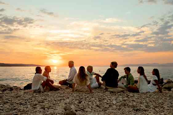 En större grupp människor sitter på en stenstrand i solnedgången.