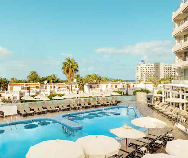 Udsigt over hotelpool med liggestole og parasoller, omgivet af bygninger og palmer. (AI-genereret tekst)