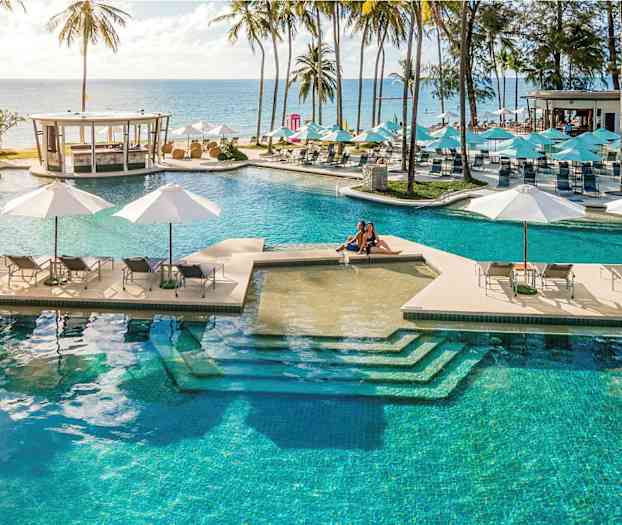 En stor pool med liggestole og parasoller med udsigt over havet, omgivet af palmer. (AI-genereret tekst)