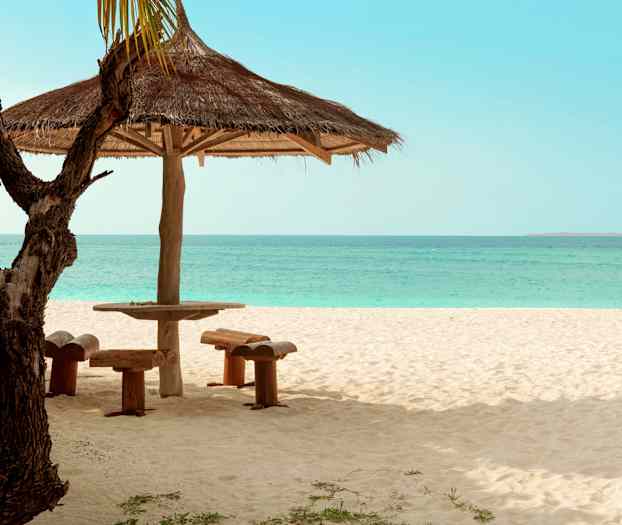 En parasol med træbord og siddepladser på en sandstrand med havet i baggrunden. (AI-genereret tekst)