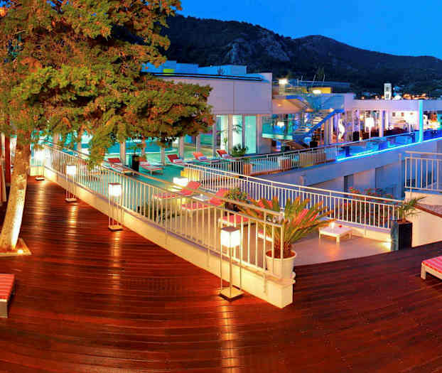 Et udendørsmiljø med træterrasse, liggestole og grønne planter nær en pool. I baggrunden ses bygninger og et bjergrigt landskab. (AI-genereret tekst)