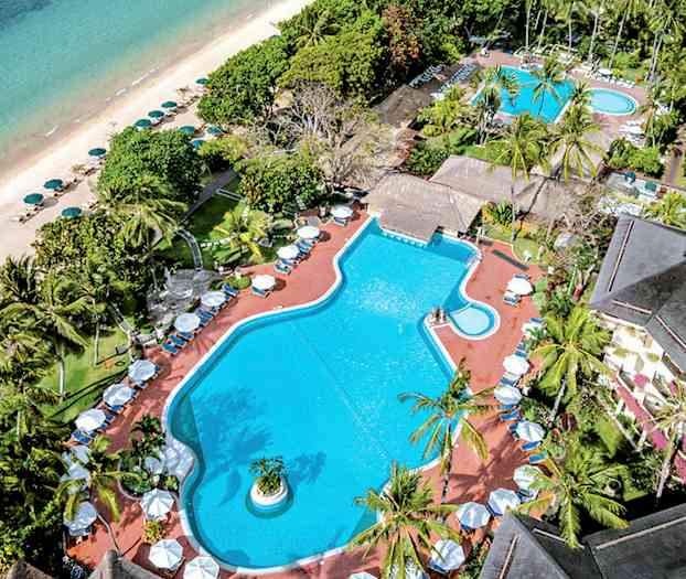 Luftfoto af et poolområde omgivet af tropiske træer, med en strand og havet i baggrunden. (AI-genereret tekst)