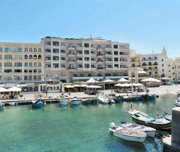 Et billede af en havn med både i klart vand foran en række bygninger med udendørs serveringsområder og terrasser. (AI-genereret tekst)