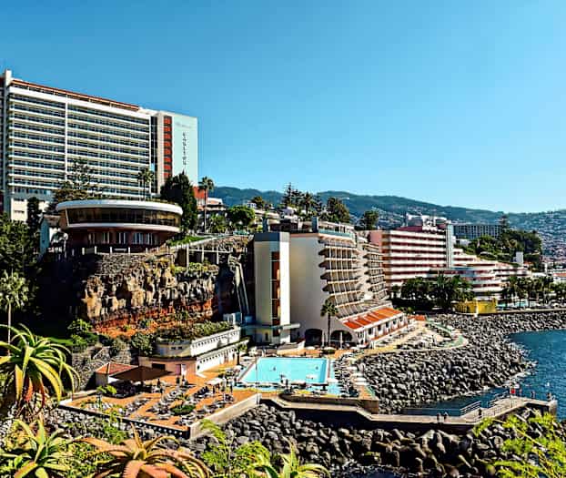 Billede af et hotel beliggende ved en kyst med udsigt over havet. Bygningen har en pool og grønne omgivelser. (AI-genereret tekst)