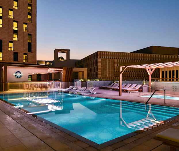 En oplyst udendørs swimmingpool om aftenen omgivet af liggestole og moderne bygningsarkitektur. (AI-genereret tekst)