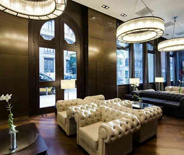 En elegant stue med en siddegruppe i Chesterfield-stil, store vinduer, lysekroner og trægulv. (AI-genereret tekst)