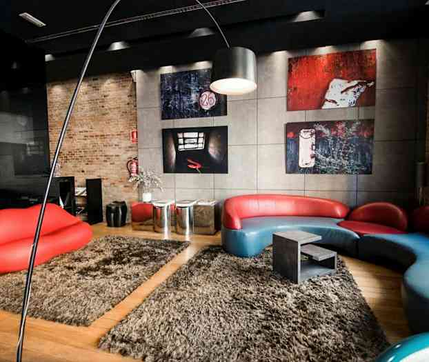 Et moderne rum med en rød og blå sofa i unikt design, en rød sofa-lignende siddemøbel formet som læber, og kunstværker på væggene. Gulvet er dækket af beige tæpper. (AI-genereret tekst)