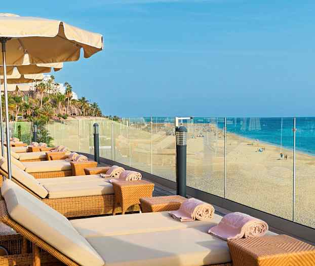 Liggestole med parasoller på en terrasse med udsigt til en sandstrand og havet. (AI-genereret tekst)