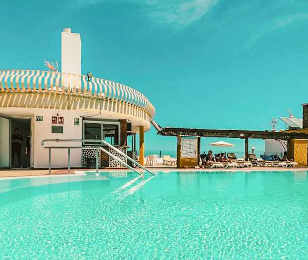 En udendørs swimmingpool ved en bygning med liggestole og en klar blå himmel. (AI-genereret tekst)