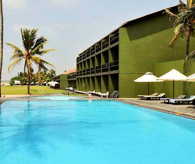 En udendørs pool ved et hotel med grønne bygninger, omgivet af palmer og liggestole under parasoller. (AI-genereret tekst)
