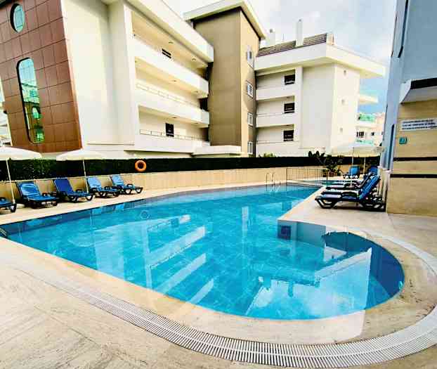 Udendørs pool med blåt vand omgivet af liggestole og bygninger. (AI-genereret tekst)