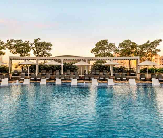 Udendørs swimmingpool omgivet af liggestole og parasoller med træer i baggrunden. (AI-genereret tekst)