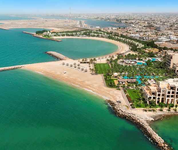 Et luftfoto af en kystby med en halvcirkelformet strand, bygninger, palmer og turkis havvand. (AI-genereret tekst)