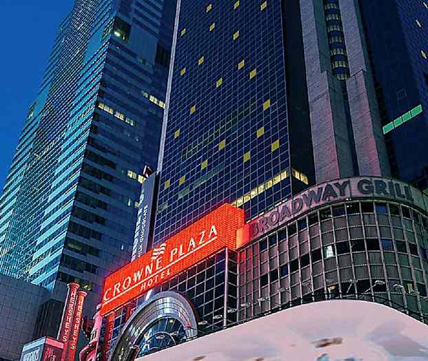 Et billede af en høj bygning med neonskilte, der viser "Crowne Plaza" og "Broadway Grill". (AI-genereret tekst)