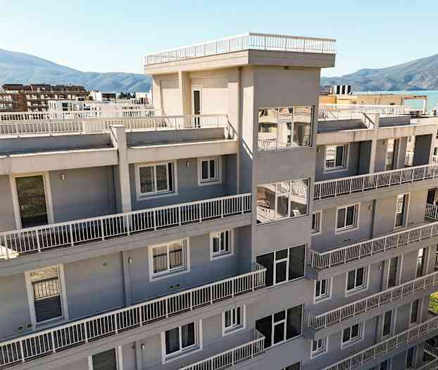 En moderne bygning med flere etager og altaner, omgivet af grønt og nærliggende bjerge i baggrunden. (AI-genereret tekst)