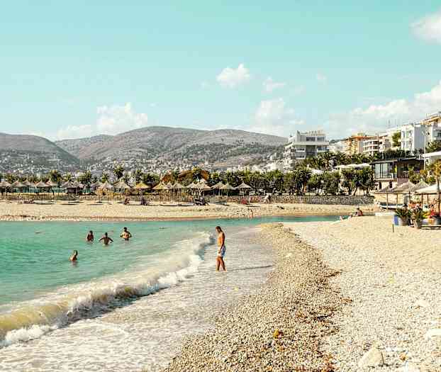 Strand med parasoller og badegæster i klart vand. Bygninger og bjerge i baggrunden. (AI-genereret tekst)