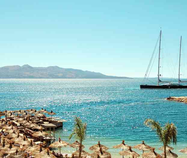 Strand med parasoller og lang sejlbåd på havet med bjerge i baggrunden. (AI-genereret tekst)