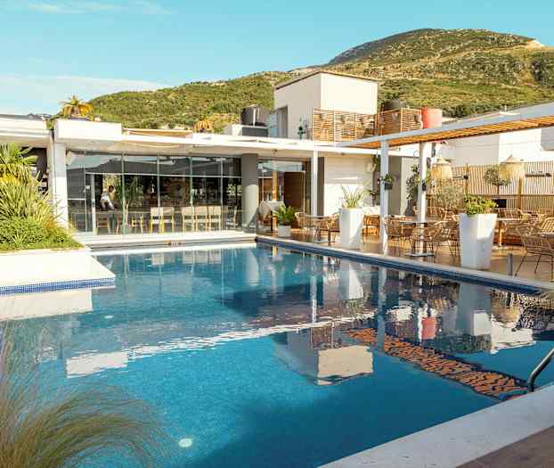 En moderne bygning med glasfacade ved siden af en udendørs pool, omgivet af grøn vegetation. (AI-genereret tekst)