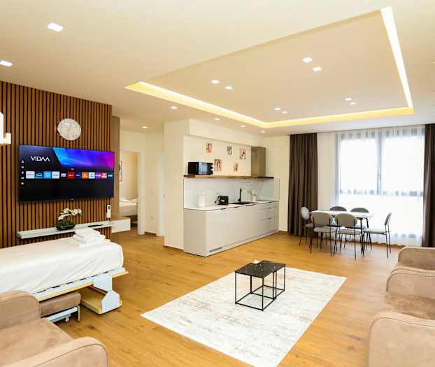 Et moderne hotelværelse med åbent gulvplan, der inkluderer en sovesofa, TV, køkkenområde, spisebord og store vinduer med gardiner. (AI-genereret tekst)