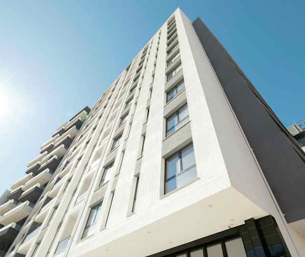 Billedet viser en moderne bygning med flere etager, facaden er lys og har mange vinduer. En klar blå himmel og direkte sollys er synlige. (AI-genereret tekst)