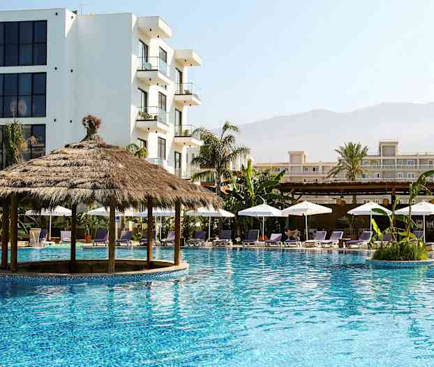 En pool med en stråtagspavillon omgivet af liggestole og parasoller, med flere bygninger i baggrunden. (AI-genereret tekst)