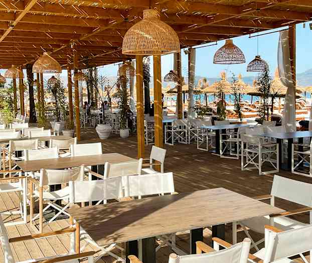 Billede af en udendørs spiseplads med træborde og hvide stole under en træpergola. (AI-genereret tekst)