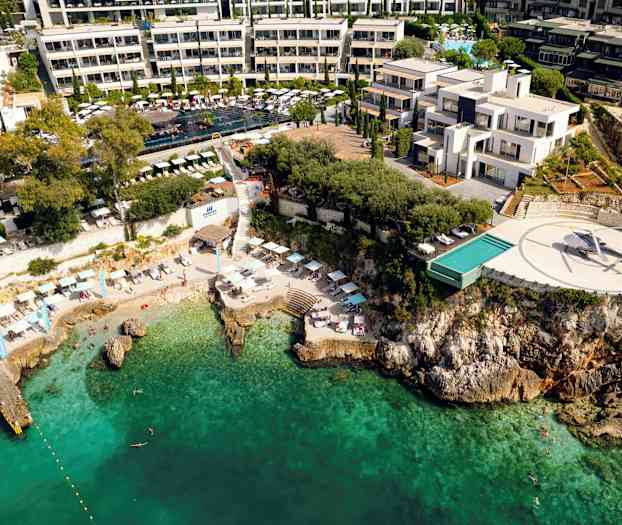 Luftfoto af en kystnær hotel med pool, helikopterlandingsplads og strand ved turkis vand. (AI-genereret tekst)