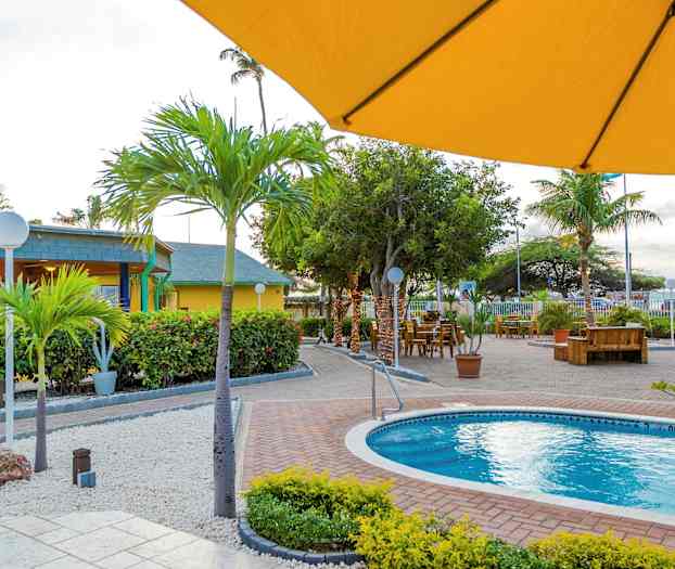 Et udendørsområde med en pool omgivet af planter, palmer og udendørsmøbler. I forgrunden ses en gul parasol. (AI-genereret tekst)