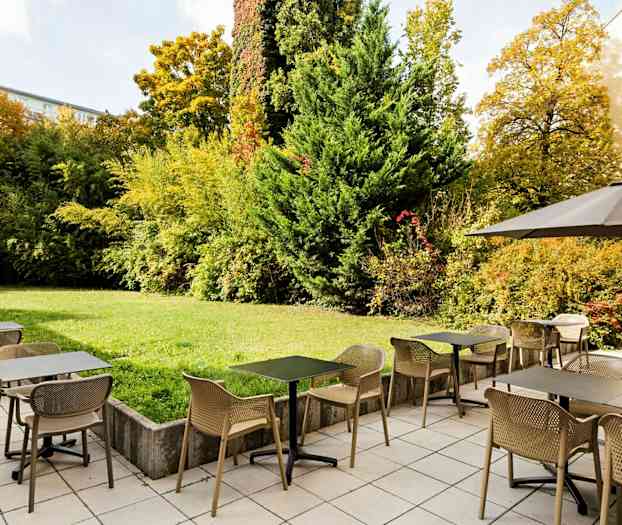 En udendørs restaurant med borde og stole på et flisebelagt område ved siden af en grøn have med buske og træer. (AI-genereret tekst)