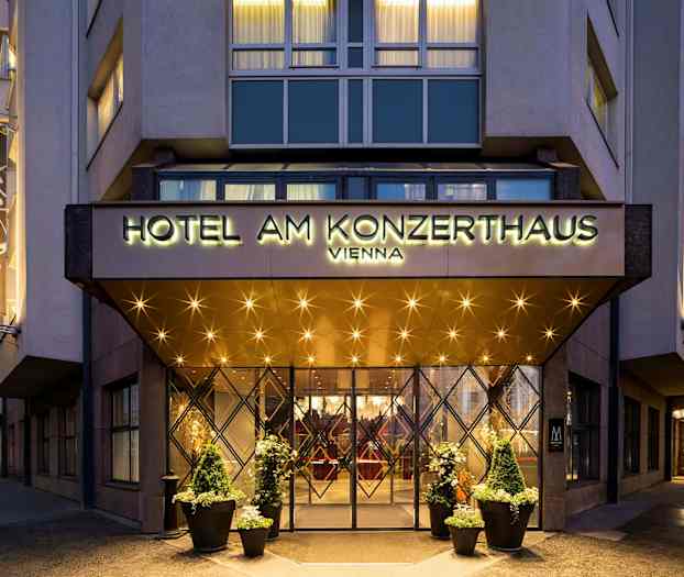 Indgangen til et hotel med et skilt, der lyder "Hotel am Konzerthaus Vienna". Belysning og dekorationer vises omkring indgangen og på facaden. (AI-genereret tekst)