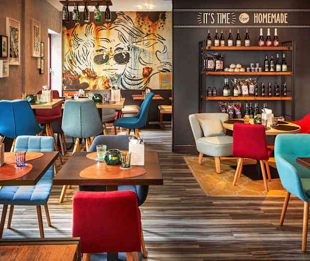 En restaurantinteriør med farverige siddemøbler, dekorative borde med drikkevarer og bestik samt en væg med hylder fyldt med forskellige produkter. (AI-genereret tekst)