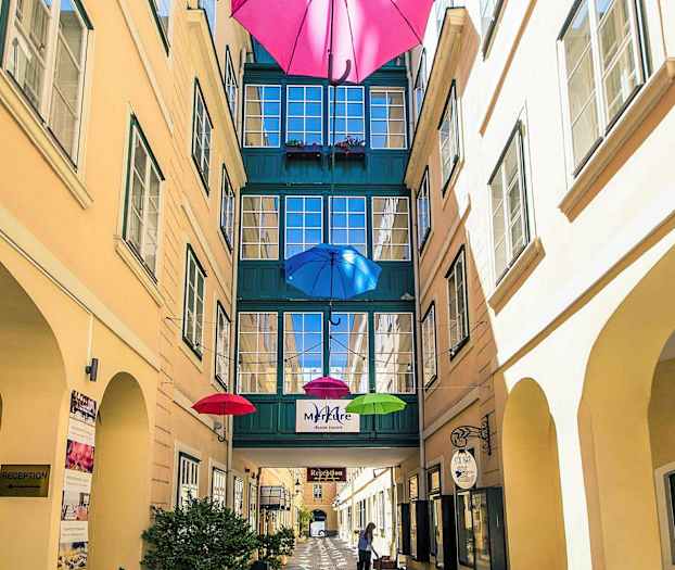 En smal gårdsplads med farverige paraplyer hængende ovenover og en passage mellem bygninger. (AI-genereret tekst)