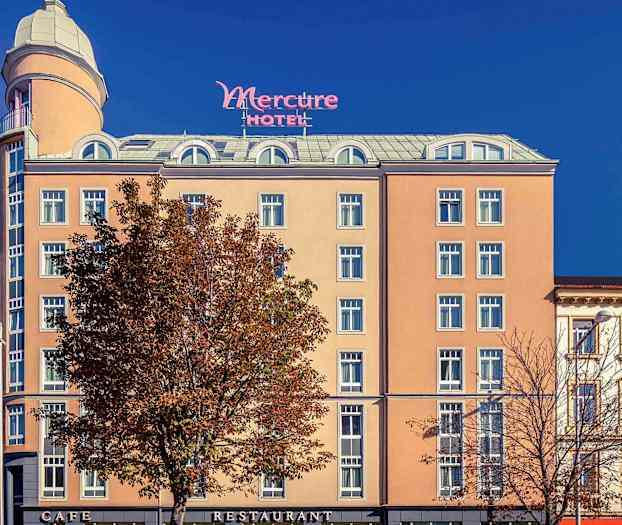 Eksteriøret af et hotel med navnet Mercure, med en beige facade, mange vinduer og et højt tårn øverst. (AI-genereret tekst)