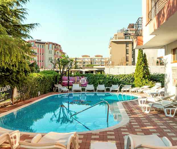 En udendørs pool omgivet af liggestole og grønne områder med bygninger i baggrunden. (AI-genereret tekst)