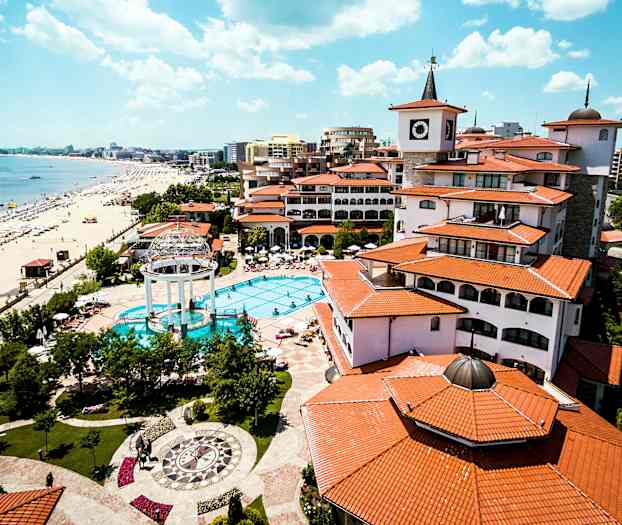 Et feriested med pool, omgivet af bygninger med røde tage, ligger ved en strand med parasoller og liggestole. (AI-genereret tekst)