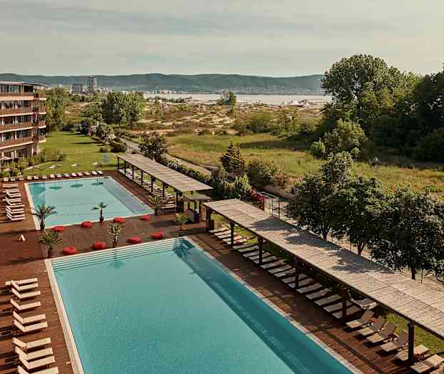 Stor pool med liggestole og parasoller ved siden af et hotel, med grønt område og landskab i baggrunden. (AI-genereret tekst)