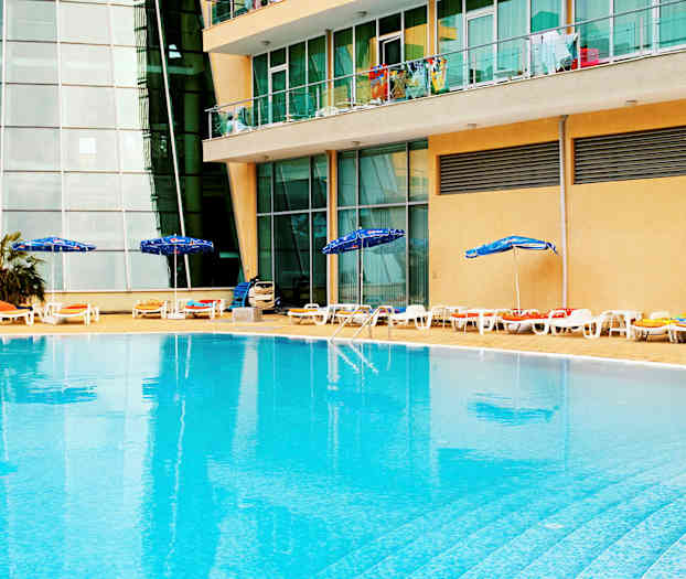En udendørs pool med liggestole og parasoller ved kanten, beliggende foran en bygning med glasvinduer og altaner. (AI-genereret tekst)