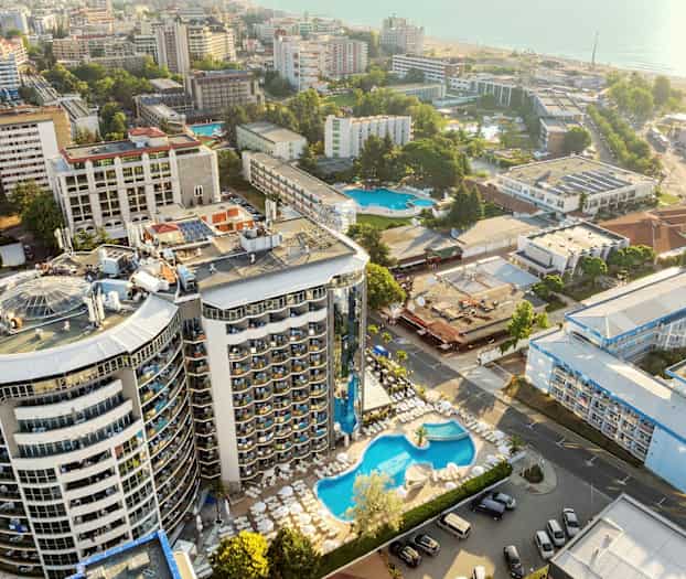 Luftfoto af hotelområde med pool og omkringliggende bygninger tæt på havet. (AI-genereret tekst)