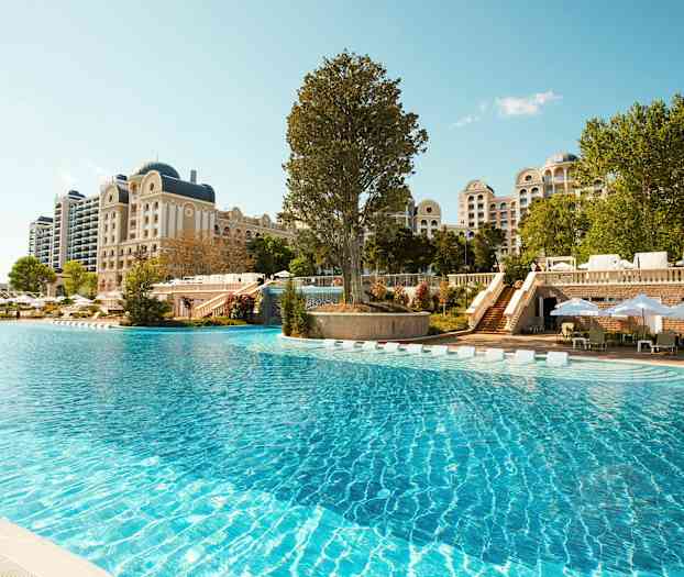 Stor udendørs pool foran et hotel med klassisk arkitektur. (AI-genereret tekst)