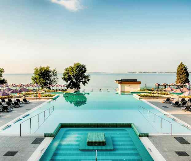 En udsigt over en pool med liggestole og parasoller spredt rundt omkring, samt havet og træer i baggrunden. (AI-genereret tekst)