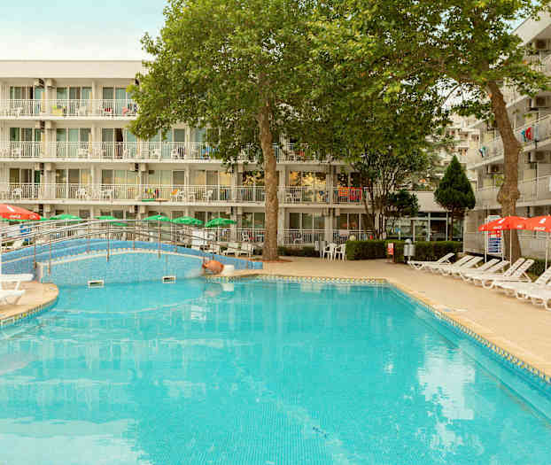 En udendørs pool med liggestole og parasoller foran en bygning med altaner. (AI-genereret tekst)