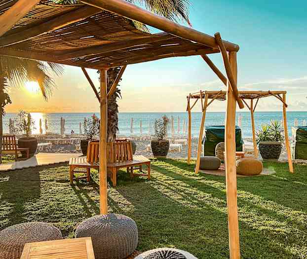 Udendørsmøbler under træskærme nær stranden ved solnedgang. (AI-genereret tekst)