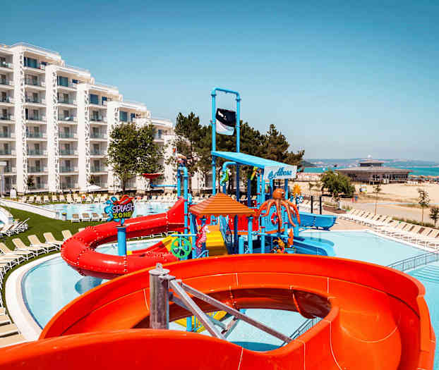 Billede af et hotelområde med pool, vandrutsjebaner og liggestole tæt på stranden. (AI-genereret tekst)