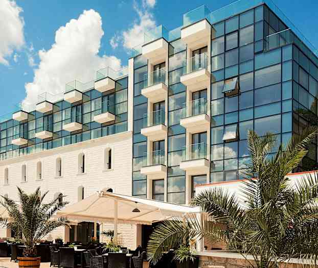 Et moderne hotel i glas og sten med altaner. Foran bygningen er der en udendørs servering med palmer. (AI-genereret tekst)