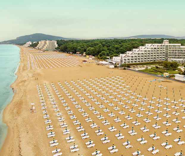 Luftfoto af en stor sandstrand med rækker af liggestole og parasoller samt bygninger og beplantning i baggrunden. (AI-genereret tekst)